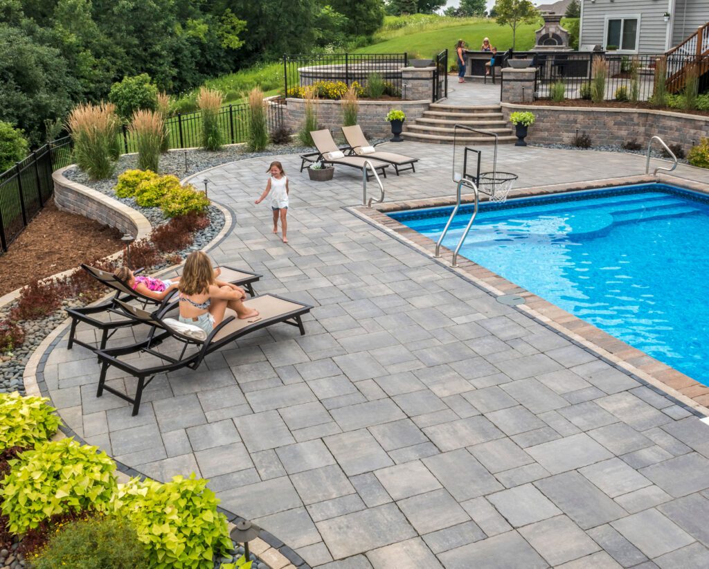 children on patio with chairs, pool with basketball hoops, and masonry landscaping